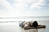 Young man lying on wet sand on beach, hands behind head