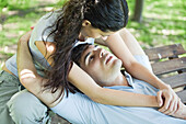 Young couple, man reclining on bench while woman puts arms around him from behind