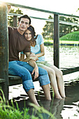 Couple sitting on dock, dangling legs in water, woman looking at camera, full length