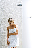 Woman standing by shower, wrapped in towel, looking down