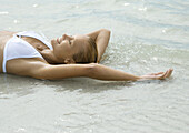 Woman lying in shallow water on beach, smiling with eyes closed