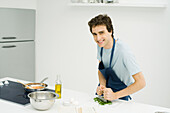 Young man mincing basil in the kitchen, smiling at camera