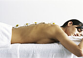 Woman lying on stomach, eyes closed, with daisies aligned on bare back, side view