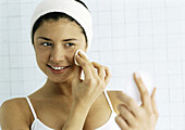 Young woman wiping face with cotton pad, looking at compact