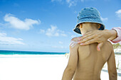 Female hand applying sunscreen to boy's bare back on beach