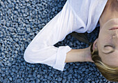 Woman lying on gravel with eyes closed and hands behind head, cropped