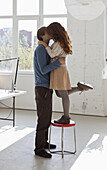 A young woman on a stool kissing her tall boyfriend