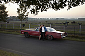 A rockabilly couple leaning against a vintage car in the country