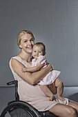 Portrait of happy woman with baby girl in wheelchair against gray wall
