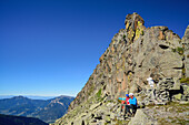 Two hikers sitting beneath a rock spire and looking at the mountains, Trans-Lagorai, Lagorai range, Dolomites, UNESCO World Heritage Site Dolomites, Trentino, Italy