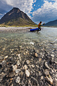 Paddler carries her pack raft across a shallow section of the Marsh Fork of the Canning river in the Arctic National Wildlife Refuge, Brooks range mountains, Alaska.