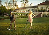 A smart young man hits a croquet ball with a mallet, a beautiful lady watches him from a distance with a mallet in her one hand and legs cross.