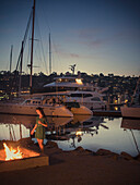 A beautiful girl with a drink in her left hand stands next to a fireplace by the marina.