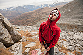 Climber wearing helmet and red jacket belaying his partner at the base of a wall in Devero National Park, with the valley in background. Ossola, Italy.