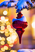 Close up of red Christmas ornament at night hanging on a snow covered evergreen tree with blurred Christmas lights in the background