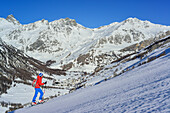 Woman back-country skiing ascending towards Punta Tre Chiosis, view to Val Varaita with Chianale, in background Tete des Toilliers, Rocca Bianca and Pic de Caramantan, Punta Tre Chiosis, Valle Varaita, Cottian Alps, Piedmont, Italy