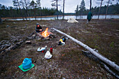 A young woman wearing a down jacket and hat sitting in front of a camp fire in a sparse forest, in front of her bivouac material, behind her a tent and a lake, Urho Kekkonen National Park, Finnish Lapland, Finland