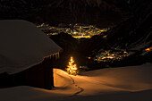 Ein beleuchteter Weihnachtsbaum vor einem Stall auf der Belalp, unten im Tal die Lichter des Dorfs Blatten und der Stadt Brig, Walliser Alpen, Kanton Wallis, Schweiz