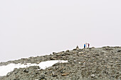 A wooden cross near the Tracuit Hut standing right where the signal for mobile phones is best, two visitors of the hut are standing next to the cross looking at their mobile phones, Val d‘Anniviers, Pennine Alps, canton of Valais, Switzerland