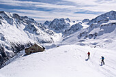 Zwei junge Skitourengängerinnen erreichen über eine weiße Kuppe die Cabane des Aiguilles Rouges oberhalb von Arolla, im Hintergrund in der Bildmitte der Mont Collon, am rechten Bildrand die Pigne d‘Arolla, Val d‘Hérens, Walliser Alpen, Kanton Wallis, Sch
