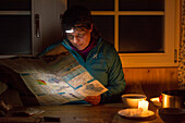 A woman with a headlight sitting at a table and looking at the skitouring map in candlelight, Cadlimo Hut, valley called Val Camadra, Lepontine Alps, canton of Ticino, Switzerland