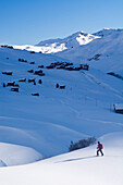 A backcountry skier walking through the snow-covered high valley called Fondei, behind him the houses of the hamlet Strassberg, Grison Alps, canton of Grison, Switzerland