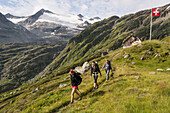 Three young women hiking along a hiking trail, behind them the Gauli Hut and a Swiss flag blowing in the wind, Gauli Region, Bernese Alps, canton of Bern, Switzerland