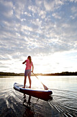 Frau beim Stand Up Paddling auf dem Chiemsee im Sonnenuntergang, Chiemgau, Bayern, Deutschland