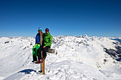 Two men sitting on the summit cross of the Schwarzhorn (3147 m), in the background on the right-hand side Piz Linard (3410 m), Grisons, Switzerland, Europe