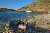 Junger Mann liegt in heißer Quelle am Strand einer einsamen Bucht auf der griechischen Insel Kithnos. Im Hintergrund liegt eine ankernde Segelyacht. Kolona, Ägäis, Kykladen, Griechenland