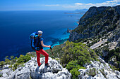 A young woman hiking along the mountainous coast, rock-needle near Pedra Longa and Santa Maria Navarrese in the background, Punta Giradili, Selvaggio Blu, Sardinia, Italy, Europe