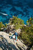 Frau klettert eine steile Wand über dem Meer hinauf, während ein Motorboot, Yacht vorbei fährt, Golfo di Orosei, Selvaggio Blu, Sardinien, Italien, Europa
