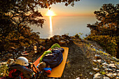 Junge Frau schläft in Schlafsack auf einem Köhlerkreis über dem Meer, bei Sonnenaufgang nahe der Bucht Cala Biriola, Golfo di Orosei, Trekking- und Kletterausrüstung, Selvaggio Blu, Sardinien, Italien, Europa