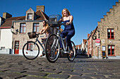 Zwei junge Frauen fahren Fahrrad auf einer Straße mit Pflastersteinen nahe der Coupure Marina, Brügge, Flandern, Belgien, Europa