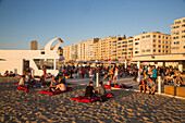 Junge Menschen chillen an einer Strandbar am Strand mit Apartments an der Strandpromenade im Hintergrund, Ostende, Flandern, Belgien, Europa