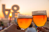 Hands hold two glasses of Belgian beer in front of Love sculpture on the beach at sunset, Ostend, Flanders, Flemish Region, Belgium