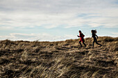 Two Young Woman Running Across Grassy Dune at Beach