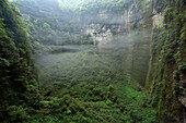 View from a small window in the wall of the vast Niubizi Tian Keng in the Er Wang Dong cave system. Three tiny cave explorers are minute in size as they wander around the heavily vegetated floor. Thin whisps of slouds gather and gently rise up following a
