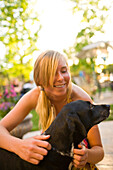 A young girl with blond pig tails pets a black dog at a barbecue on a sunny day.