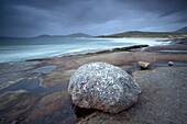 Large erratic boulder on the rocky coastline at Scarista with a view over the sand towards the hills of Taransay, Scarista, Isle of Harris, Outer Hebrides, Scotland, United Kingdom, Europe