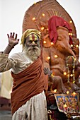 Holy man raises a hand in front of a Ganesh statue draped in fairy lights at the Hindu festival of Shivaratri, Pashupatinath, Kathmandu, Nepal, Asia