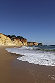 A wave breaks on golden sands flanked by steep cliffs, typical of the Atlantic coastline near Lagos, Algarve, Portugal, Europe