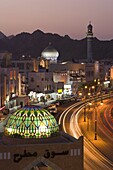 Elevated dusk view along the Corniche, stained glass dome of Mutrah Souq, colourful latticed buildings and Mutrah Mosque, Muscat, Oman, Middle East