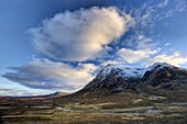 Winter view of Rannoch Moor showing lone whitewashed cottage on the bank of a river, dwarfed by snow-covered mountains, and dramatic evening sky, Rannoch Moor, near Fort William, Highland, Scotland, United Kingdom, Europe
