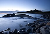 The ruins of Dunstanburgh Castle at dawn with Greymare Rock partly sumberged and the sea blurred by a long exposure, Embleton Bay, Northumberland, England, United Kingdom, Europe