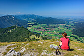 Woman hiking sitting at Gamsknogel and looking towards Inzell, Chiemgau Alps in background, Gamsknogel, Chiemgau Alps, Upper Bavaria, Bavaria, Germany