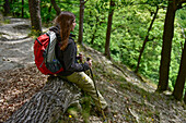 Junge Frau pausiert auf einem Baumstamm bei Wanderung durch Wald auf dem Wanderweg Harzer Hexen Stieg von Thale nach Treseburg, Frühjahr, Harzvorland, Harz, Sachsen-Anhalt, Deutschland
