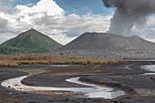 Eruption des aktiven Vulkans Tavurvur mit Aschewolke, im Vordergund Rinnsal aus Regenwasser im Aschesand, im Hintergrund die grüne Kegelspitze des ''Vulcan'', Papua Neuguinea, Neu Britannien, Süd Pazifik