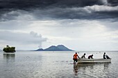 Küste von Rabaul bei Ebbe. Unterspülung einer kleinen Insel wird sichtbar. Im Hintergrund der aktive Vulkan Tarvurvur mit Eruption, im Vordergrund Männer, die ein Boot besteigen. - Rabaul, Papua Neuguinea, Neu Britannien