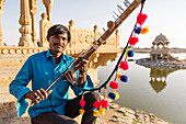 Indian man holding traditional instrument near monument, Jaisalmer, Rajasthan, India